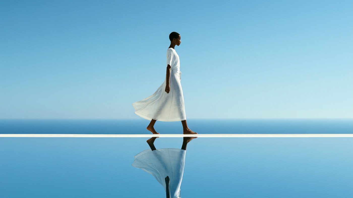 A woman in a white dress walking a mirrored ledge above a still blue sea